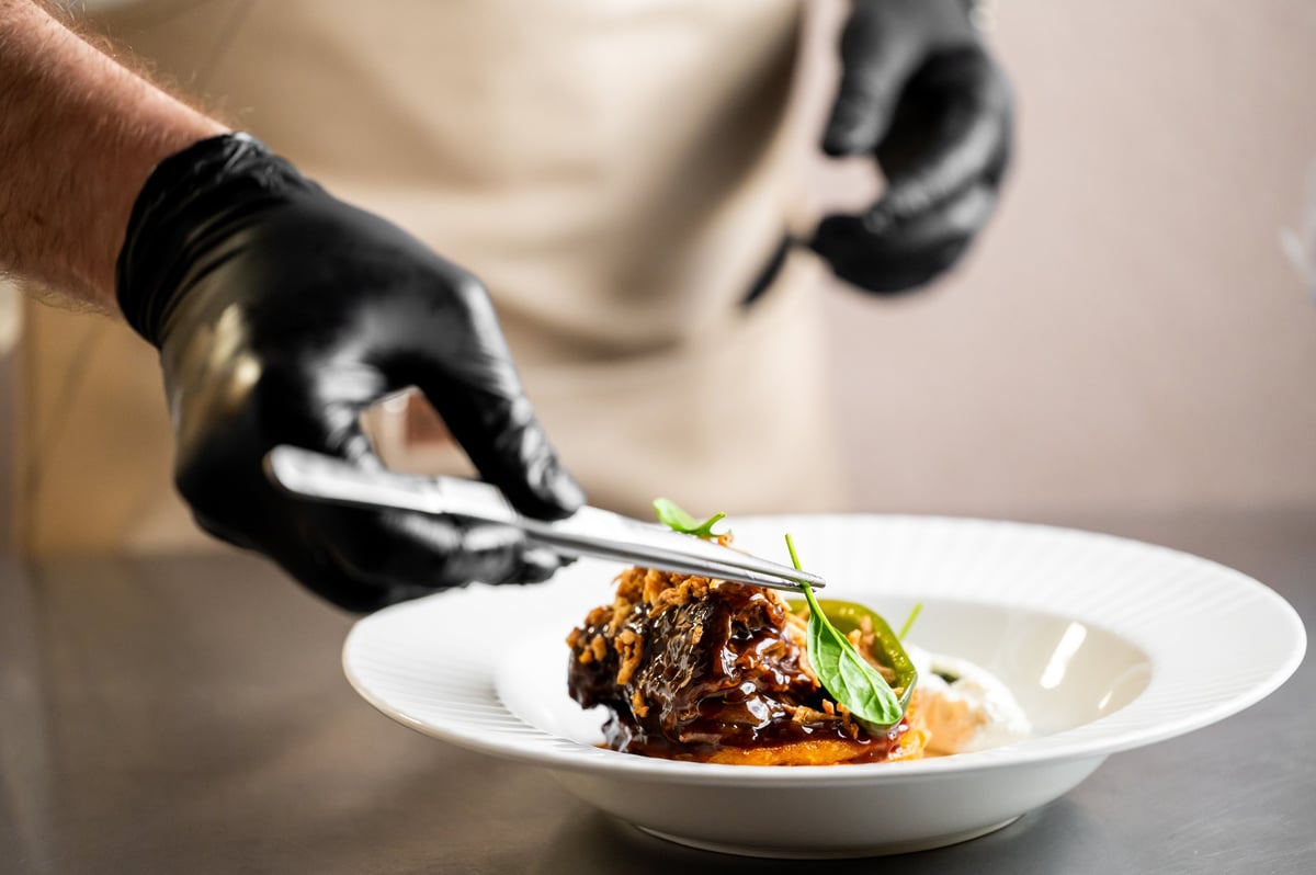 A chef in black gloves carefully placing garnish on a gourmet meat dish