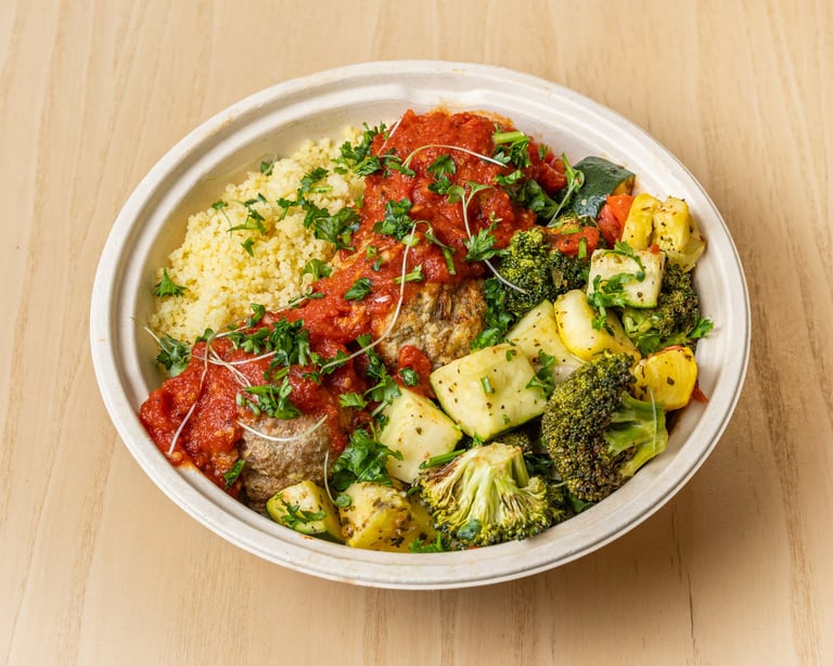 Healthy bowl meal with roasted broccoli, cauliflower, couscous, and grilled meat topped with fresh herbs and red sauce on a wooden table