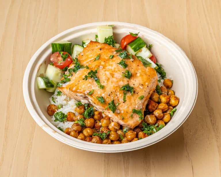Grilled salmon fillet with roasted chickpeas, cucumber, tomato, and herbs served in a white bowl on wooden surface