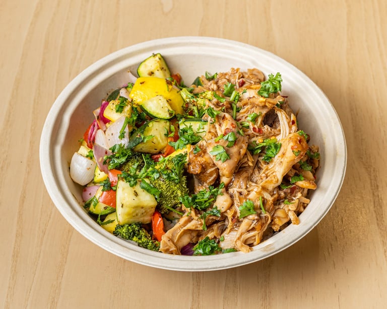 Bowl of shredded chicken and roasted potatoes with fresh herbs and vegetables on wooden surface