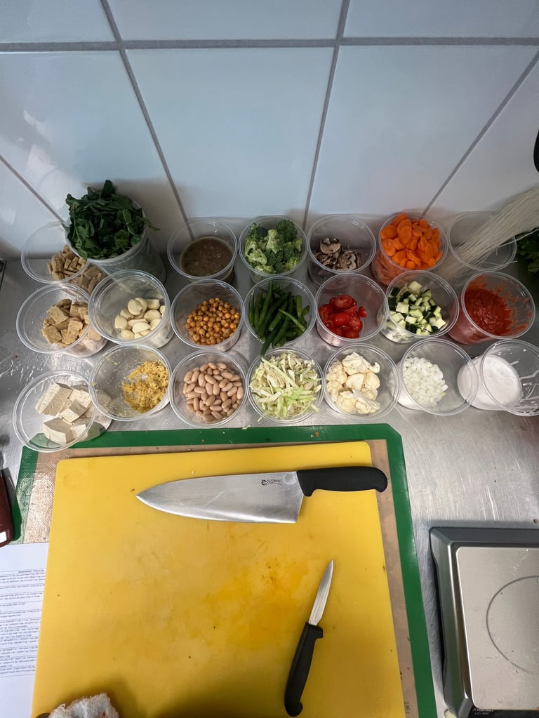 Overhead view of kitchen prep station with multiple clear containers of prepped ingredients, yellow cutting board, and chef knives