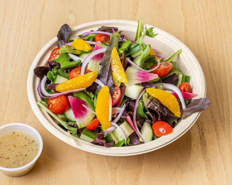 Colorful salad bowl with mixed greens, orange segments, radishes, tomatoes, and cucumber on wooden surface with dressing on side